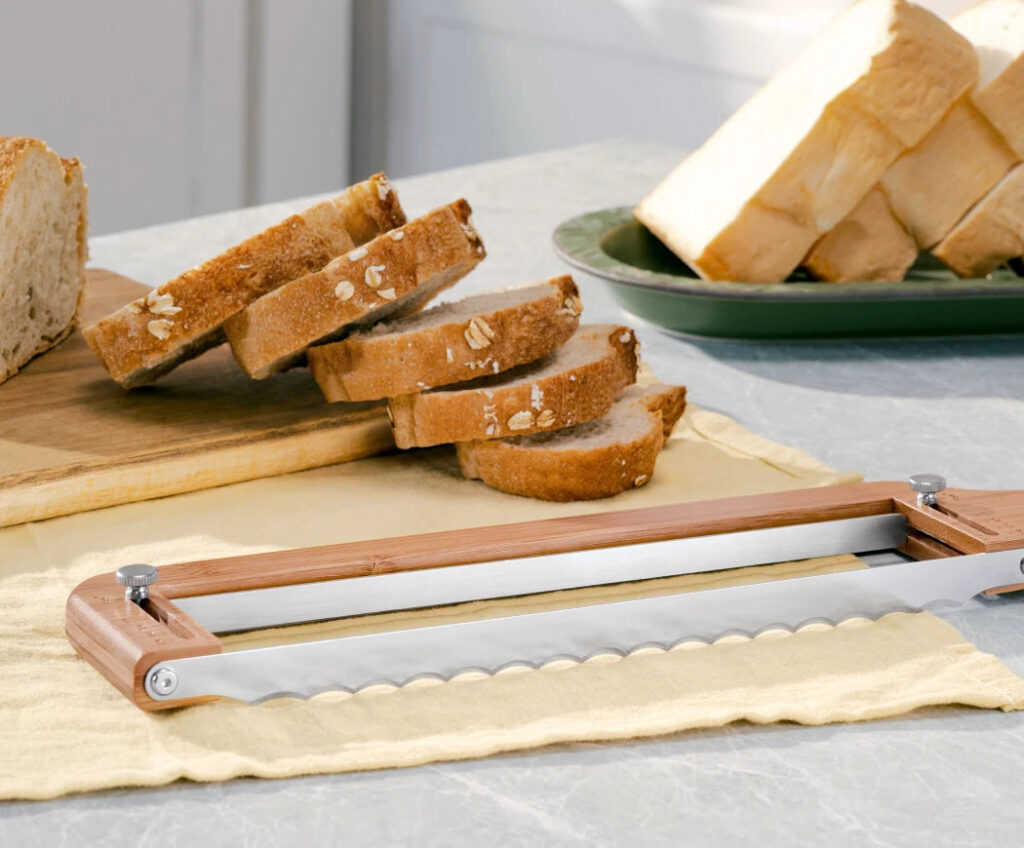How to Freeze Sourdough Bread: Whole Loaves or Slices? Bamboo adjustable bread slicer on a cloth beside two groups of sliced bread.